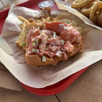 Bentley's Roast Beef - Lobster Roll - A lobster roll featuring chopped lobster meat mixed with celery, served in a toasted bun on a red tray lined with parchment paper. Accompanying the lobster roll are onion rings and a small cup of dipping sauce, with additional fried items visible in the background. The setting appears to be a casual dining environment with a wooden table and outdoor seating. - Sandwiches Near Me - Amherst, New Hampshire