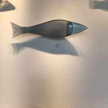 Blue Rain Gallery - Glass fish - A glass fish sculpture with a streamlined, elongated body and striped pattern is mounted on a white wall. The sculpture has a transparent head and tail, with a darker, striped midsection. It casts a shadow on the wall beneath it. The setting appears to be an art gallery, with other glass artworks partially visible in the background. - Art Galleries Near Me - Santa Fe, New Mexico