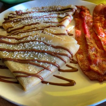 Lemon Leaf Cafe - Nutella Crepes w/ side of bacon - The image shows a white plate with two folded crepes drizzled with chocolate sauce and sprinkled with powdered sugar, placed next to three strips of cooked bacon. The crepes appear soft and are garnished with a generous amount of chocolate and sugar. The bacon is crispy and reddish-brown, positioned on the right side of the plate. The background suggests a casual dining setting, possibly at Lemon Leaf Cafe. - Breakfast & Brunch Near Me - Largo, Florida