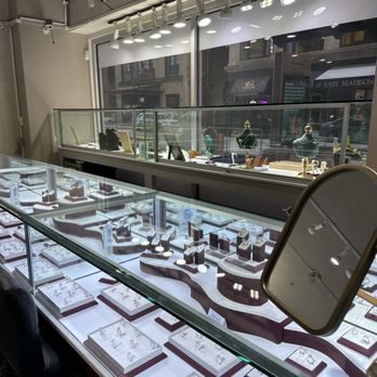Diamonds - The image shows the interior of a jewelry store with a glass display case filled with rings and necklaces, arranged on white platforms. The store has large windows with a view of the street outside, and a glass barrier separates the display from the window. Overhead track lighting provides illumination, and a mirror is positioned on the side of the display case. The scene suggests a retail environment focused on jewelry sales. - Jewelry Near Me - Chicago, Illinois