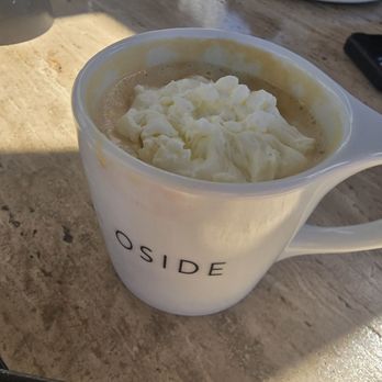 High/Low - Late - A white ceramic mug with the text 'O SIDE' printed on it holds a hot beverage topped with whipped cream. The mug is placed on a wooden table, with sunlight casting shadows nearby. In the background, there are other plates and glasses, indicating a dining or cafe setting. - Breakfast & Brunch Near Me - Oceanside, California