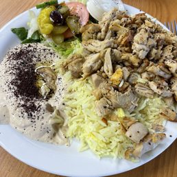 Chicken Shawarma Platter - A white plate contains a serving of yellow rice, chopped cooked chicken, a side salad with lettuce, tomato, cucumber, and pickles, and a dollop of creamy white sauce topped with black pepper. The plate is placed on a wooden table, with a small container of additional sauce and a fork visible in the background.