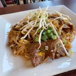 Pad Thai - A plate of Pad Thai featuring stir-fried noodles topped with bean sprouts and chopped green onions. The dish is served on a white square plate placed on a wooden table in a restaurant setting, with chairs and tables visible in the background.