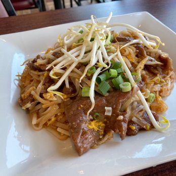 Manee Thai Restaurant - Pad Thai - A plate of Pad Thai featuring stir-fried noodles topped with bean sprouts and chopped green onions. The dish is served on a white square plate placed on a wooden table in a restaurant setting, with chairs and tables visible in the background. - Thai Near Me - Gray, Maine