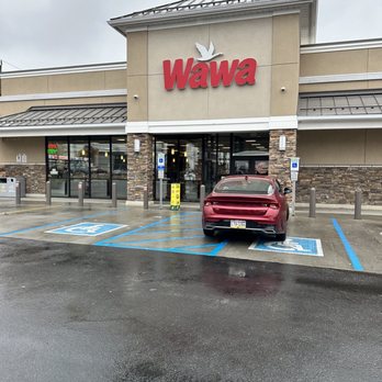 Wawa - The image shows the exterior of a Wawa convenience store with a large red Wawa sign and dove logo above the entrance. The storefront has glass doors and windows, with stone accents on the lower part of the building. In front, there are designated accessible parking spaces, one of which is occupied by a red car. The parking lot appears wet, and the sky is overcast, indicating rainy weather. - Gas Stations Near Me - Maple Shade, New Jersey