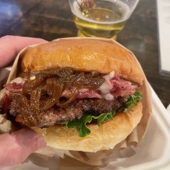 Rustic Nomads - A burger with caramelized onions, meat patty, lettuce, and toppings, served in a paper wrapper with a glass of beer behind it. - Food Trucks Near Me