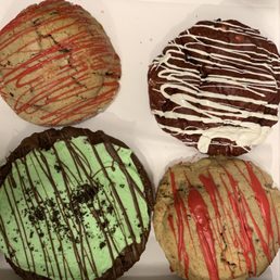 The image shows four large cookies arranged on a white tray. Each cookie has a distinct appearance: one with green icing and chocolate drizzle, another with red icing and red drizzle, a third with chocolate icing and chocolate drizzle, and the last with a brownish base and white drizzle. The cookies are decorated with various toppings and icing patterns, highlighting their size and flavor variety.