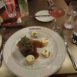 Filet mignon and La Vie en Rose cocktail - A restaurant table features a large white plate with 'MONARQUE' printed on the rim, holding a filet mignon topped with greens, four dollops of mashed potatoes garnished with chives, and a brown sauce. Surrounding the plate are two glasses of water, a pink cocktail in a coupe glass, and two tall glasses with mint leaves and lime slices. The setting suggests a fine dining experience with elegant presentation and a variety of beverages.