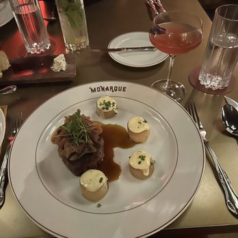 Monarque - Filet mignon and La Vie en Rose cocktail - A restaurant table features a large white plate with 'MONARQUE' printed on the rim, holding a filet mignon topped with greens, four dollops of mashed potatoes garnished with chives, and a brown sauce. Surrounding the plate are two glasses of water, a pink cocktail in a coupe glass, and two tall glasses with mint leaves and lime slices. The setting suggests a fine dining experience with elegant presentation and a variety of beverages. - French Near Me - Baltimore, Maryland