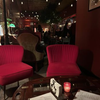 CODE RED - Inside - The image shows a restaurant interior with two pink upholstered chairs in the foreground, a dark wooden table with menus and a small red candle holder. In the background, there are more tables, chairs, and a bar area with patrons and staff. The ceiling has numerous hanging light bulbs, creating a warm, dim ambiance. A large tree or plant is visible in the middle of the space, adding to the decor. The overall setting appears intimate and stylish. - Cocktail Bars Near Me - Washington, DC, District of Columbia
