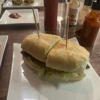 310 Eatery - The Crispy Chicken Temescal Sandwich - A sandwich featuring a bread roll, lettuce, and a meat patty is placed on a white square plate. The sandwich is cut in half and held together with toothpicks. In the background, there are various condiments, sauces, and other dishes, including small burgers with skewers. The setting appears to be a restaurant or casual dining environment. - Burgers Near Me - Albany, California