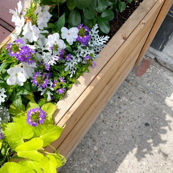 Chadwick's - A wooden planter box contains various flowers, including white, purple, and yellow blooms, along with green leaves, placed on a concrete surface. - Canadian (New) Near Me - Toronto, Ontario
