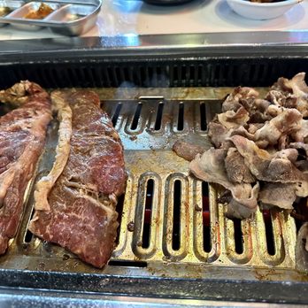Meat Project - Cooking meat. - Thin slices of beef and pork are placed on a tabletop grill, with some meat partially cooked and others raw. The grill is embedded in the table, typical of Korean BBQ restaurants. In the background, small dishes and bowls with side items are visible, indicating a communal dining setting focused on grilling meat. - Korean Near Me - Centreville, Virginia