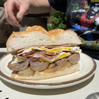 Caffe Social - A sandwich featuring sausage, fried eggs, cheese, and sliced bread is presented on a white plate outdoors. The background includes a person wearing a black graphic T-shirt with colorful designs, and greenery is visible. The sandwich appears freshly made, with the ingredients layered inside a soft bread roll. - American Near Me - Norwalk, Connecticut