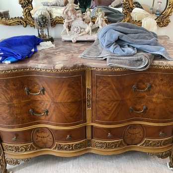 XS Trash - A wooden dresser with ornate carvings and a marble top, topped with towels, a clock, and decorative figurines, with a mirror above. - Junk Removal & Hauling Near Me - Pompano Beach, Florida