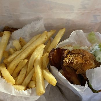 Birdcall - All Natural Chicken - A takeout container holds a serving of French fries and a fried chicken sandwich. The fries are golden and crispy, placed in a paper cup. The fried chicken sandwich includes a breaded chicken fillet, lettuce, and bacon, wrapped in paper. The box is open, revealing the food inside, with a paper liner at the bottom. - Chicken Shop Near Me - Denver, Colorado