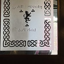 A sign with Celtic knot border, featuring a lion emblem and the text 'Maggie Miley's Irish Pub' in reverse, seen through a window.