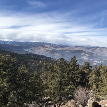 Gregory Canyon - The image captures a panoramic view of a mountainous landscape with a foreground of green pine trees and rocky terrain. Distant mountain ranges with some snow-capped peaks are visible under a partly cloudy sky with streaks of white clouds. The scene suggests a high-altitude, natural environment with clear visibility and varied terrain. - Hiking Near Me - Boulder, Colorado