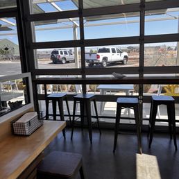 The image shows the interior of a coffee shop with a large glass window that provides a view of parked vehicles outside, including a white pickup truck and a gray SUV. Inside, there is a wooden counter with a tissue box and a small mirror, along with several black barstools positioned along a high counter facing the window. The space is illuminated by natural light, and outdoor tables and chairs are visible through the window.