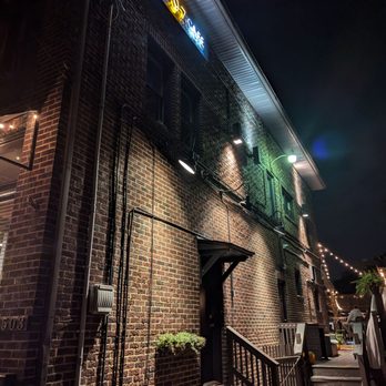 Old 97 Cafe - Outside - The image shows the exterior of a brick building at night, featuring several exterior lights that illuminate the brick wall. A neon sign displaying 'Old 97 Cafe' is mounted near the roofline. The building has multiple windows, a small staircase leading to a door, and string lights visible in the background, suggesting an outdoor patio area. - Jazz & Blues Near Me - Akron, Ohio