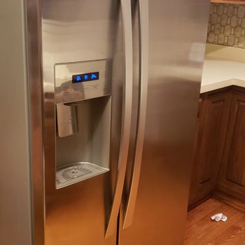 ACM Movers - A stainless steel side-by-side refrigerator featuring a water and ice dispenser on the left door, with digital controls above the dispenser. The refrigerator is positioned in a kitchen with wooden cabinets and hardwood flooring, and a small piece of paper or cloth is visible on the floor nearby. - Movers Near Me - Evanston, Illinois
