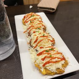 Tostones - A rectangular white plate holds multiple sushi rolls topped with mayonnaise, ketchup, and small pieces of chopped green onions. The rolls are placed on a dark wooden table, with a glass of water, a folded napkin, and a person's arm visible in the background. The presentation includes shredded cheese or crab on top of the rolls, with a drizzle of sauces for flavor.