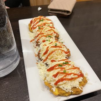 Bienmesabe - Tostones - A rectangular white plate holds multiple sushi rolls topped with mayonnaise, ketchup, and small pieces of chopped green onions. The rolls are placed on a dark wooden table, with a glass of water, a folded napkin, and a person's arm visible in the background. The presentation includes shredded cheese or crab on top of the rolls, with a drizzle of sauces for flavor. - Venezuelan Near Me - Chicago, Illinois