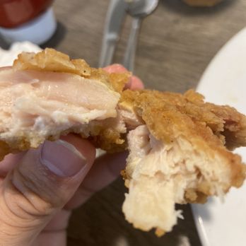 Buttered Toast Restaurant - Undercooked chicken tender kids meal - A person holds a fried chicken tender with a golden-brown, crispy coating. The tender has a bite taken out, revealing white meat inside. The background shows a wooden table with utensils, a glass of yellow beverage, and condiments, indicating a restaurant setting. - Breakfast & Brunch Near Me - Algonquin, Illinois