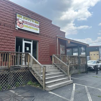 Big Fatty's BBQ - Outside - The photo depicts the front exterior of Big Fatty's BBQ restaurant, with a brick building facade and a sign displaying the restaurant's name and contact information. A wooden ramp with stairs provides access to the entrance, which has a glass door. The parking lot in front contains multiple parked vehicles, and there is a small round table with a metal surface near the ramp. The sky is partly cloudy, and neighboring industrial buildings are visible in the background. - Barbeque Near Me - White River Junction, Vermont