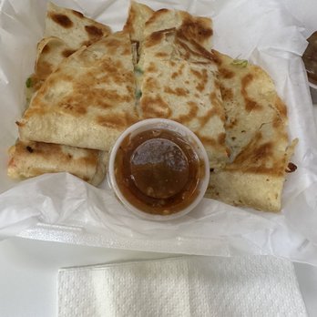 Papa Nachos - A serving of quesadillas, cut into triangular pieces, is presented on a white paper-lined tray. The quesadillas are golden brown with some darker spots, indicating they are toasted. A small clear cup of dark dipping sauce, possibly salsa or gravy, is placed in the center of the tray. The presentation is casual, typical of a Mexican or New Mexican cuisine setting. - Mexican Near Me - Albuquerque, New Mexico