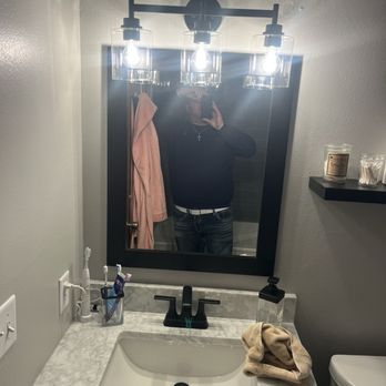 Construction Specialties - A person is capturing a mirror selfie in a bathroom, with a modern light fixture above the mirror featuring three exposed bulbs. The bathroom has a marble countertop with a sink, toothbrush holder, soap dispenser, and a towel. A shelf on the wall holds candles and cotton swabs. The person is wearing a dark top, jeans, and a cross necklace, with their face partially obscured by the phone. - Decks & Railing Near Me