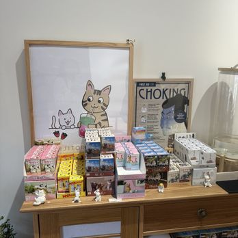 Kijitora E14th - Cute cat display inside - The image shows a wooden table with multiple small animal figurines arranged in front of colorful boxes. Behind the table, there are two illustrated posters: one features a cat and a dog, and the other depicts a person with a sign about choking. The setting appears to be inside a shop or café, with a plain wall background and some additional items partially visible on the right. - Coffee & Tea Near Me - New York, New York
