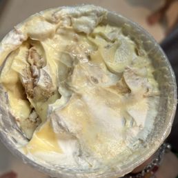 Banana Pudding - A cup of banana pudding with a creamy, smooth texture, containing visible slices of banana and possibly cookie or wafer pieces. The pudding appears thick and well-mixed, with a slightly glossy surface. The image is taken from above, showing the dessert's surface and ingredients clearly.