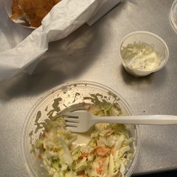 Coleslaw - A clear plastic bowl contains partially eaten coleslaw made of shredded cabbage and carrots, with a white plastic fork resting inside. Next to it is a small cup of tartar sauce. In the background, there is a basket lined with white paper holding fried fish pieces. The items are placed on a metallic surface, likely a table at a casual dining or takeout setting.