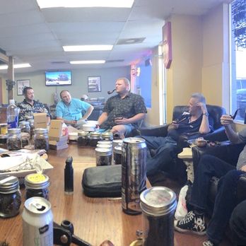 Cigar Inn - A group of people are seated around a large rectangular table inside a room, engaging in conversation or using devices. The table has various jars, cans, and personal items. Some individuals are holding cameras or phones, and a window in the background shows parked cars outside. The room has a casual atmosphere with shelves and a television on the wall. - Tobacco Shops Near Me - Phoenix, Arizona