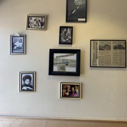 Decor - The image shows a beige wall decorated with several framed photographs and newspaper clippings. The frames vary in size and color, including black, silver, and gold. The photographs include black-and-white and color images of people, some in vintage attire, and one appears to be a newspaper article from the Glendale Republic. The arrangement is informal, with frames placed at different heights. The lower part of the wall has a wooden wainscot paneling.