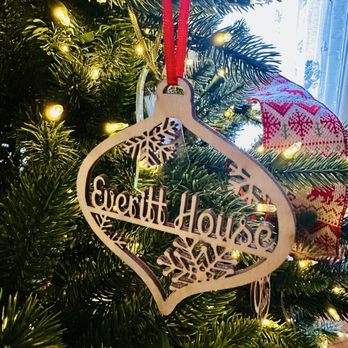 Everitt House - A wooden Christmas ornament with the words "Everitt House" cut out hangs on a decorated Christmas tree. The tree is adorned with small yellow lights and a red ribbon. A red and white patterned stocking is visible in the background near a window with sheer curtains. - Bed & Breakfast Near Me - Hackettstown, New Jersey