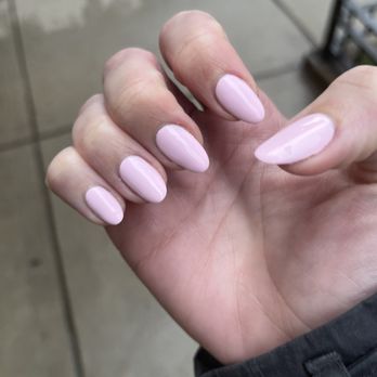 Nail 2ne1 Spa - No-chip mani - The image features a close-up of a person's hand displaying five fingers with freshly painted, pale pink, almond-shaped nails. The background shows a sidewalk and part of an outdoor setting, with a blurred metal object visible in the upper right corner. The person is wearing a dark-colored sleeve or jacket cuff. - Nail Salons Near Me - Chicago, Illinois