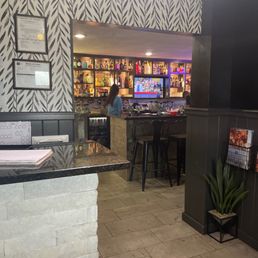 Bar - A bar with shelves of liquor bottles, a television, and a person working behind the counter, with patterned wallpaper and bar stools.