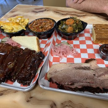 Backyard Barbeque - Ribs, Brisket, corn bread, baked beans, collard greens - A meal featuring barbecue ribs, sliced brisket, baked beans, collard greens, potato salad, cornbread, and pickled onions on a checkered paper tray. - Barbeque Near Me - Freeport, New York