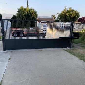 VG Tech - Newly installed automated gate by VG tech - A black metal automated sliding gate with electronic control panel is installed at a residential driveway, with trees and houses visible in the background. - Fences & Gates Near Me - Ontario, California