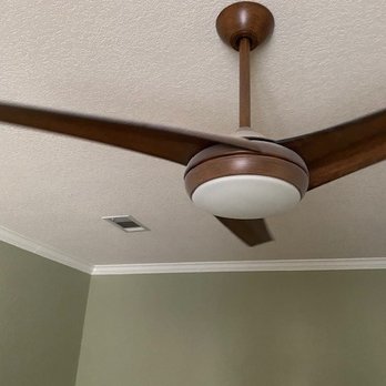 Texas Cleaning Services - The image shows a ceiling fan with three wooden blades and a central light fixture mounted on a textured ceiling. The fan is attached to a short downrod connected to a round ceiling mount. The ceiling has a textured finish, and part of a green wall with white crown molding is visible. A small rectangular vent is also seen on the ceiling. - Home Cleaning Near Me - Austin, Texas