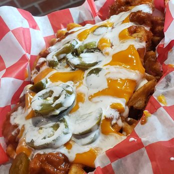 AJ's - Loaded Fries - A close-up of a basket of loaded fries topped with melted cheese, sliced jalapeños, and drizzled with sauce. The fries are served in a red and white checkered paper-lined basket, with a brick wall visible in the background. The dish appears to be a savory snack or appetizer, featuring toppings commonly associated with American-style comfort food. - American Near Me - West Lafayette, Indiana