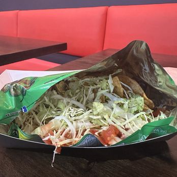 Antojitos - Todtiverduros - A salad with shredded lettuce, chopped tomatoes, and fried food items is placed in a black and green wrapper on a dark wooden table. The background features a red cushioned bench and a pink wall with yellow circular patterns, indicating a casual dining environment. - Mexican Near Me - Tucson, Arizona