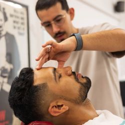 Beardbrand Barbershop - Beardbrand barber, Cisco, precisely lining up a client's hairline while the client reclines in a vintage red barber chair.