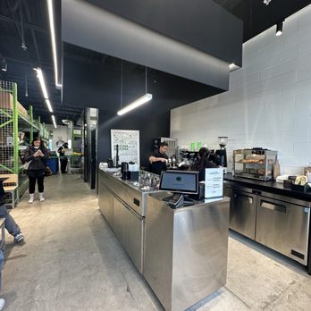 Matcha Matcha - Interior - The image shows the interior of a coffee shop with a minimalist design, including stainless steel counters and equipment. Baristas are preparing beverages behind the counter, while customers are visible in the background, some waiting and some using their phones. The ceiling is black with modern lighting fixtures, and the walls are white with a textured brick pattern. Shelving with products is visible on the left side of the image. - Coffee & Tea Near Me - Markham, Ontario