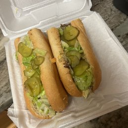 California Cheesesteak Sandwich - Two cheesesteak sandwiches with lettuce and sliced pickles are placed in a white foam takeout container. The sandwiches are made with hoagie rolls and contain beef, lettuce, and pickles. The container is open, revealing the sandwiches, and is set on a marble countertop. The background includes part of a kitchen or food preparation area.