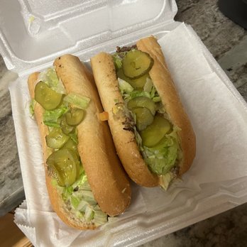 Crossroads Hotel - California Cheesesteak Sandwich - Two cheesesteak sandwiches with lettuce and sliced pickles are placed in a white foam takeout container. The sandwiches are made with hoagie rolls and contain beef, lettuce, and pickles. The container is open, revealing the sandwiches, and is set on a marble countertop. The background includes part of a kitchen or food preparation area. - Pizza Near Me - Hellertown, Pennsylvania