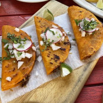 Mexico Authentico - Birrias tacos - Three tacos with a golden, slightly charred corn tortilla shell are arranged on a wooden serving board lined with parchment paper. Each taco is topped with sliced radishes, chopped onions, and cilantro. Lime wedges are placed beside the tacos for added flavor. The background shows additional dishes and utensils, indicating a dining setting at a Mexican restaurant emphasizing traditional flavors and fresh ingredients. - Mexican Near Me - Brooklyn, New York