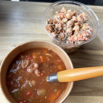 Electric Beets - Bean soup and chickpea salad - The image shows a disposable paper cup filled with bean soup containing various vegetables and beans, with a yellow spoon inside. Next to it is a clear plastic container filled with chickpea salad mixed with vegetables and dressing. The items are on a light-colored wooden table, with a blurred background suggesting an outdoor or casual dining setting. - Vegan Near Me - Brooklyn, New York